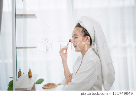 Woman Applying Skincare Routine at Home with Towels Wrapped Around Hair in Bright Modern Bathroom with Green Plants and Natural Light 131749049