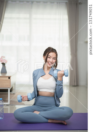 Young woman in activewear cheerfully exercising at home with dumbbells, promoting fitness and healthy lifestyle while sitting on yoga mat 131749051