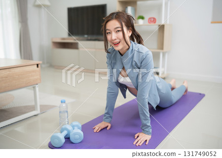Young woman in activewear practicing yoga on mat at home, using dumbbells, focusing on fitness and wellness, healthy lifestyle and exercise routine 131749052
