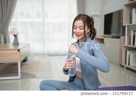 Young woman in active wear opening water bottle while practicing yoga in bright modern living room with natural light and minimalistic decor Young woman in active wear opening water bottle while practicing yoga in bright modern living room with natural light and minimalistic decor 131749117