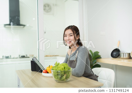 Young woman smiling while preparing healthy meal in modern kitchen with fresh vegetables and tablet on the table for recipe guidance Young woman smiling while preparing healthy meal in modern kitchen with fresh vegetables and tablet on the table for recipe guidance 131749124