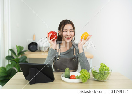 Young woman in casual kitchen holds red and yellow bell peppers while smiling, preparing healthy meal with fresh vegetables and tablet on table 131749125