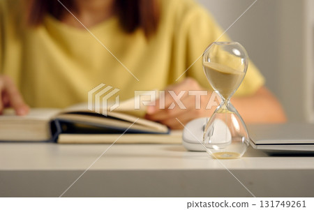 Glass hourglass standing against background of woman reading a book closeup. Glass hourglass standing against background of woman reading a book closeup. 131749261