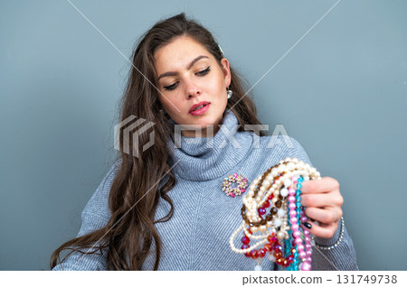 elegant woman in a long gray sweater has fun with many bead chains and jewelry, isolated on gray 131749738