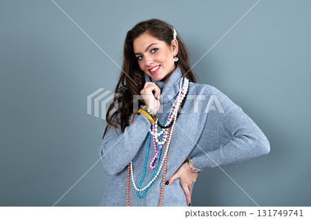 elegant woman in a long gray sweater has fun with many bead chains and jewelry, isolated on gray 131749741