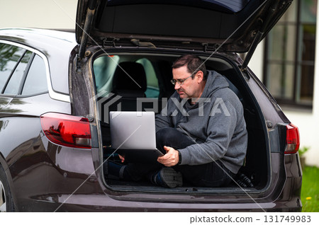 a man sitting in the open trunk of a car and working with a laptop, mobile technology, remote work 131749983