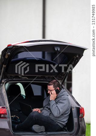a man sitting in the open trunk of a car and working with a laptop, mobile technology, remote work 131749985