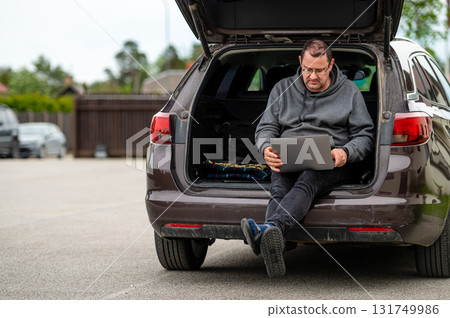 a man sitting in the open trunk of a car and working with a laptop, mobile technology, remote work 131749986