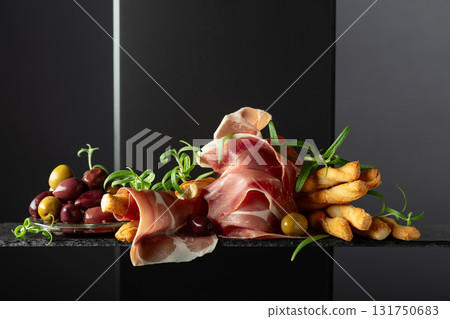 Prosciutto with bread sticks, olives, and rosemary on a black background. 131750683