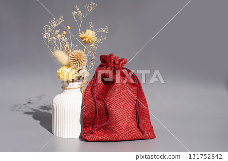 Red drawstring bag with dried flowers in ceramic vase on gray background Red drawstring bag with dried flowers in ceramic vase on gray background 131752842