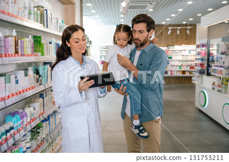 Smiling pharmacist talking with man and child giving advice about product in bright drugstore Smiling pharmacist talking with man and child giving advice about product in bright drugstore 131753211