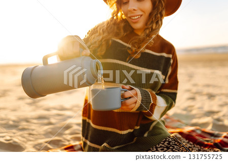 Young woman sits at picnic on the beach drinks a hot drink from a thermos. Relax and lifestyle Young woman sits at picnic on the beach drinks a hot drink from a thermos. Relax and lifestyle 131755725