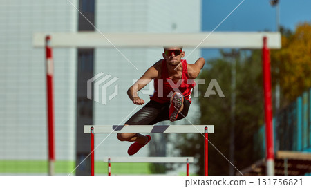 Male hurdler in powerful jump over barrier with city stadium in background Male hurdler in powerful jump over barrier with city stadium in background 131756821
