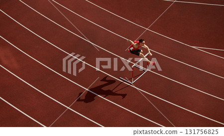 Aerial view of athlete jumping over hurdle on red running track with shadow Aerial view of athlete jumping over hurdle on red running track with shadow 131756825
