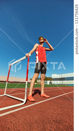 Confident male athlete resting near hurdle on running track under blue sky Confident male athlete resting near hurdle on running track under blue sky 131756826