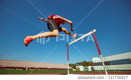 Rear view of hurdler jumping high over barrier under blue sky 131756827
