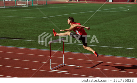 Athlete in red sprinting and jumping over hurdle on green stadium track Athlete in red sprinting and jumping over hurdle on green stadium track 131756828