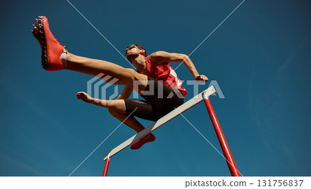 Athlete in red leaping over hurdle with power and confidence under clear sky 131756837