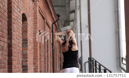 A woman performs a standing stretch, arms behind her back, leaning on a brick wall. This scene highlights the dedication to flexibility exercises and the importance of taking time for self-care. 131757711