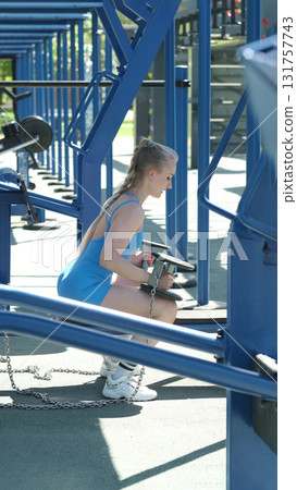 In a blue sports bodysuit, a woman lifts dumbbells at an outdoor gym, focusing on strength training exercises. The photo emphasizes the advantages of combining weight training with outdoor activities 131757743