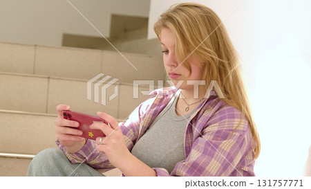 A young woman sitting on stairs smiles while looking at her phone. The image reflects casual moments of digital connection and the joy of staying in touch with loved ones. A young woman sitting on stairs smiles while looking at her phone. The image reflects casual moments of digital connection and the joy of staying in touch with loved ones. 131757771