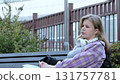 A young woman relaxes on a bench, leaning back with her arms crossed and headphones resting around her neck. The photo captures a peaceful moment of rest amidst a busy day. 131757781