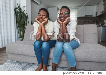 Two cheerful black twin sisters watching television sitting on sofa at home 131757889