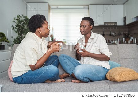 Two Young Adult Black Women Relaxing on Sofa, Drinking Tea and Chatting 131757890