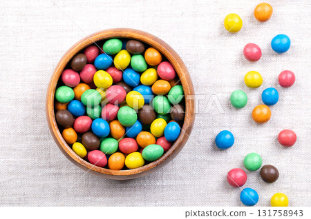 Colorful sugar-coated chocolate dragees, in a wooden bowl, on linen. Dragee chocolate confectionery, consisting of a multi colored candy shell, surrounding a chocolate filling. Close-up, from above. Colorful sugar-coated chocolate dragees, in a wooden bowl, on linen. Dragee chocolate confectionery, consisting of a multi colored candy shell, surrounding a chocolate filling. Close-up, from above. 131758943