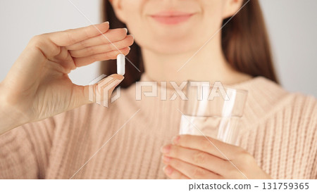 Close-up of a smiling unrecognizable woman holding a white capsule and a glass of water before taking medication or vitamin. Concept of treatment, prevention and daily healthcare routine. Close-up of a smiling unrecognizable woman holding a white capsule and a glass of water before taking medication or vitamin. Concept of treatment, prevention and daily healthcare routine. 131759365
