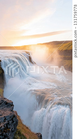 Golden sunset illuminating powerful gullfoss waterfall, cascading through misty spray with dramatic golden light spreading across rugged icelandic landscape 131759778