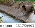 The end of an old concrete pipe of large diameter protrudes from the ground above a ditch with a flowing small river with reinforced concreted banks. Road underground communications 131759903