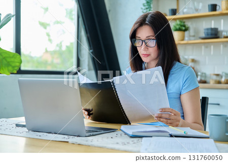 Young serious woman working with laptop, reading business papers, sitting at table at home Young serious woman working with laptop, reading business papers, sitting at table at home 131760050