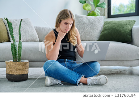 Serious focused young woman looking at laptop screen sitting on floor at home Serious focused young woman looking at laptop screen sitting on floor at home 131760194