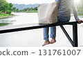A woman with a cloth bag sitting on a wooden riverfront deck gazing at the calm river, symbolizing slow living, digital detox, solo retreat, and emotional recharge in nature for self care and balance. 131761007