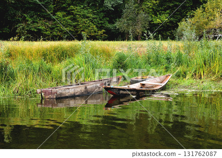 Two old boats on the shore are tied to an old car wheel, on a background of green grass a sunny summer day Two old boats on the shore are tied to an old car wheel, on a background of green grass a sunny summer day 131762087