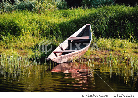 old wooden boat on the river bank close up against the background of green grass and bushes, reflections in the water 131762155