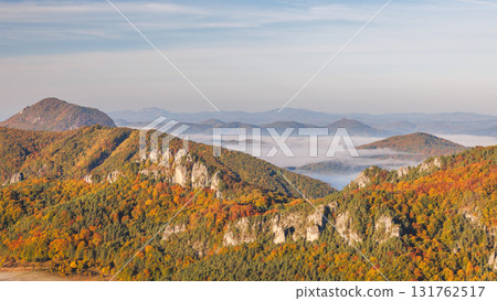 Mountains in autumn foliage, partly shrouded in mist, under a pale blue sky offer a scenic and peaceful landscape view. The Sulov Rocks, national nature reserve in northwest of Slovakia, Europe. 131762517