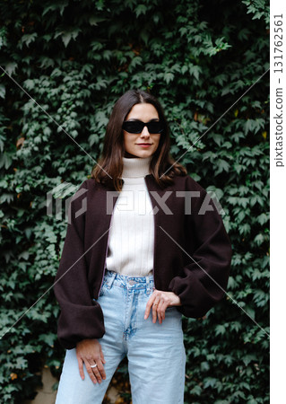 Stylish woman posing in front of green ivy wall wearing sunglasses, turtleneck sweater and bomber jacket 131762561