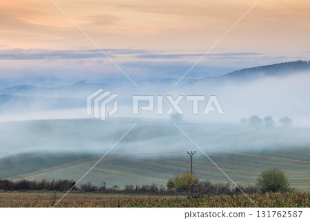 Misty landscape featuring rolling hills partially obscured by fog with trees and sky in the background. Misty landscape featuring rolling hills partially obscured by fog with trees and sky in the background. 131762587