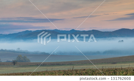 A serene vista of low-lying fog nestled between rolling hills under a pastel-tinged sky at sunrise. A serene vista of low-lying fog nestled between rolling hills under a pastel-tinged sky at sunrise. 131762608
