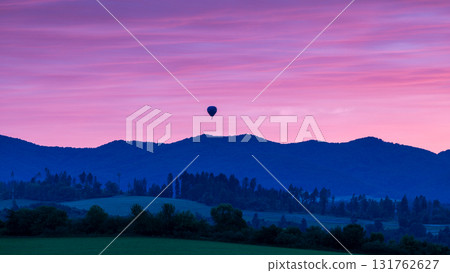 A solitary hot air balloon drifts gently across a twilight sky, framed by majestic, shadowy mountain silhouettes below. 131762627