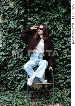 Stylish woman posing on a chair by a green ivy wall, wearing sunglasses and casual clothes Stylish woman posing on a chair by a green ivy wall, wearing sunglasses and casual clothes 131762657