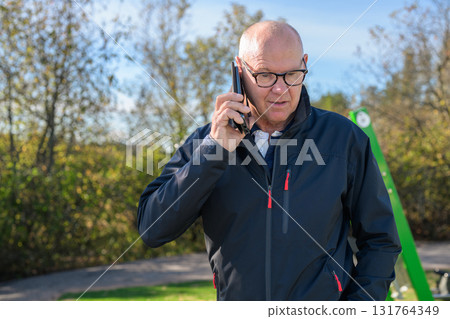 Portrait of a bald senior man talking on the phone outdoors in a park, wearing glasses and a jacket 131764349