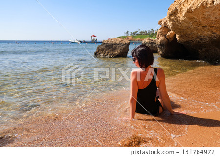 A brunette woman with glasses in a black bathing suit sits on the sand overlooking the sea, view from the back A brunette woman with glasses in a black bathing suit sits on the sand overlooking the sea, view from the back 131764972