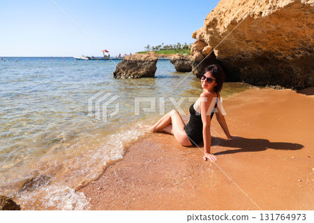 A brunette woman with glasses in a black bathing suit sits on the sand overlooking the sea, view from the back A brunette woman with glasses in a black bathing suit sits on the sand overlooking the sea, view from the back 131764973