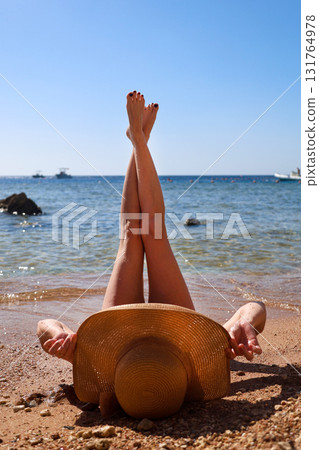 A woman in a wide sun hat lies on the sand overlooking the sea with her legs raised to the sky, enjoying the sun's rays A woman in a wide sun hat lies on the sand overlooking the sea with her legs raised to the sky, enjoying the sun's rays 131764978