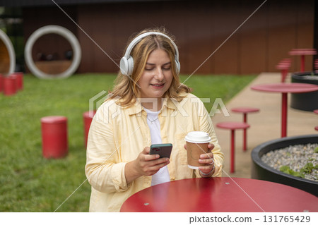 Happy young Caucasian woman in good mood listening to music with headphones and sitting on terrace street cafe in the city. Music lover enjoying music. Urban lifestyle concept. Happy young Caucasian woman in good mood listening to music with headphones and sitting on terrace street cafe in the city. Music lover enjoying music. Urban lifestyle concept. 131765429