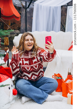 Cute young smiling woman in red sweater sitting on the floor near sofa during cozy Xmas holidays at home. Smiling Caucasian millennial lady makes selfie on phone in a Christmas decor. Full body Cute young smiling woman in red sweater sitting on the floor near sofa during cozy Xmas holidays at home. Smiling Caucasian millennial lady makes selfie on phone in a Christmas decor. Full body 131766006