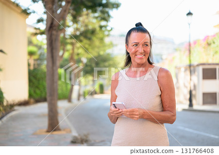 Portrait of smiling mature tourist woman using mobile phone outdoors in city park street in Spain 131766408